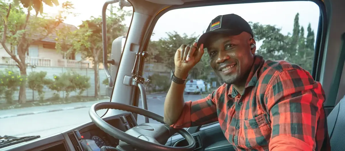 Smiling truck driver in a red plaid shirt tipping his cap from the driver's seat